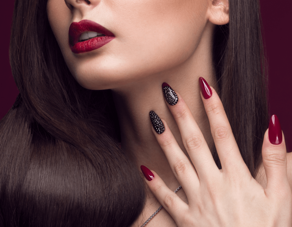 A woman's hand with sophisticated nail art featuring black and silver bead embellishments on two nails, complementing her deep red polished nails and lipstick, highlighting expert nail stylist work