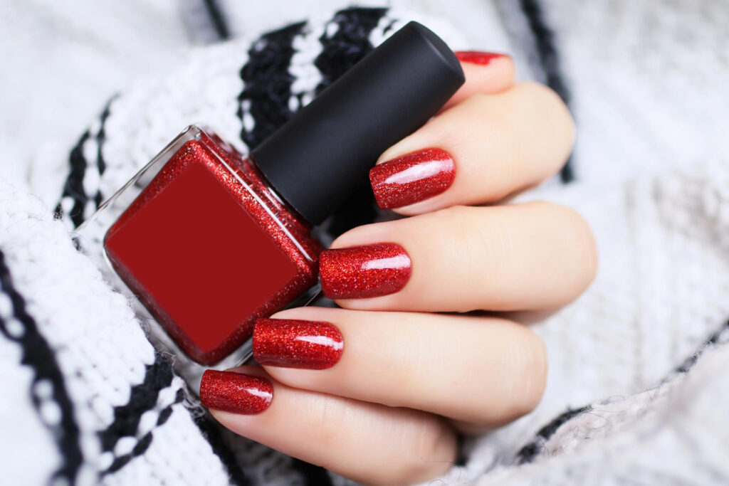 Hand holding a bottle of glittering red nail polish matching the sparkling manicure, showcasing a festive and luxurious nail stylist's work against a cozy knitted background.
