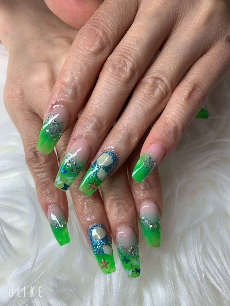 Vivid green and blue nail art with star and moon designs, featuring glitter accents, from The Grand Nails of America in Houston