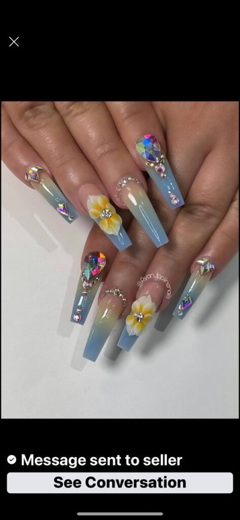 Elegant coffin-shaped nails with a blue to clear gradient, adorned with rhinestones and floral art, at The Grand Nails of America salon in Houston, Texas.