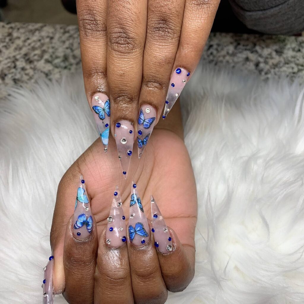 Intricate clear stiletto nails adorned with blue butterfly decals and sparkling embellishments, a signature design from The Grand Nails of America in Houston, Texas